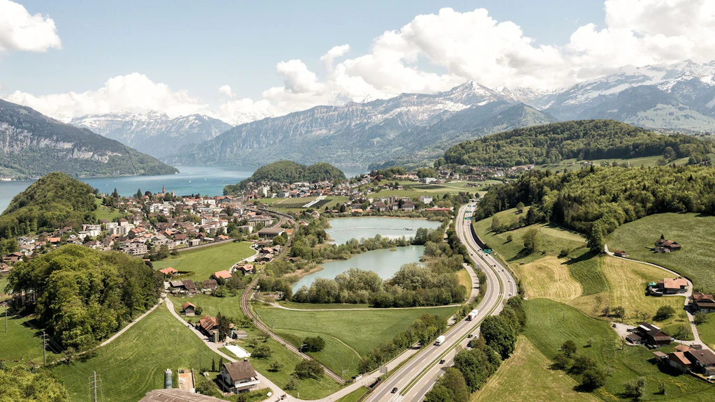 Eine Autobahn mit mehreren Fahrspuren verläuft durch eine grüne Hügellandschaft. Rechts und links der Strasse liegen Wiesen, Felder und kleine Wälder. Im Vordergrund sind einige Häuser und ein kleiner See zu sehen. Im Hintergrund erstreckt sich ein grösserer See, umgeben von Bergen mit schneebedeckten Gipfeln. Der Himmel ist mit weissen Wolken bedeckt.