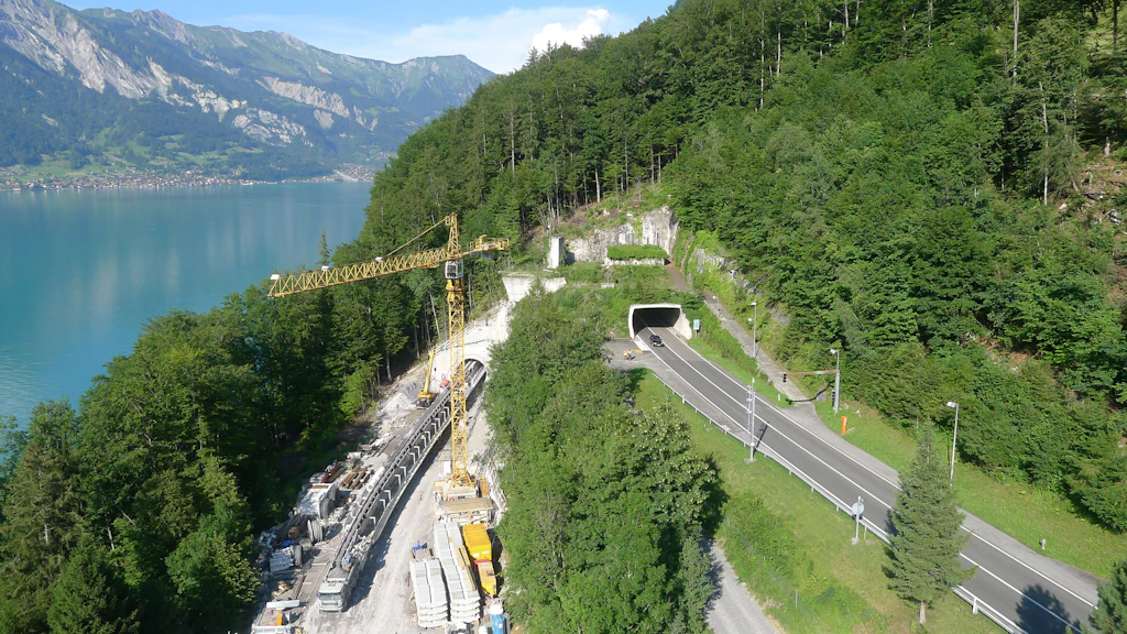 Paysage avec un lac, des montagnes, un chantier avec l'entrée d'un tunnel, une grue de chantier et une route