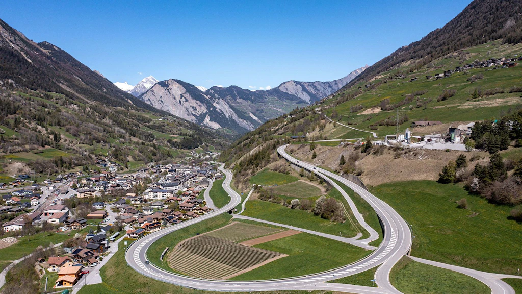Bergtal mit kurviger Strasse, landwirtschaftlichen Feldern und Dorf am Talboden, umgeben von bewaldeten und felsigen Bergen