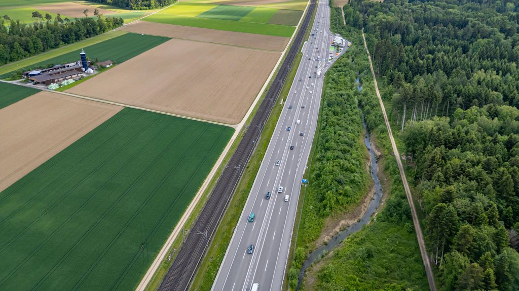 Luftaufnahme einer Autobahn mit mehreren Fahrzeugen zwischen landwirtschaftlichen Feldern und einem Waldgebiet mit einem kleinen Bach