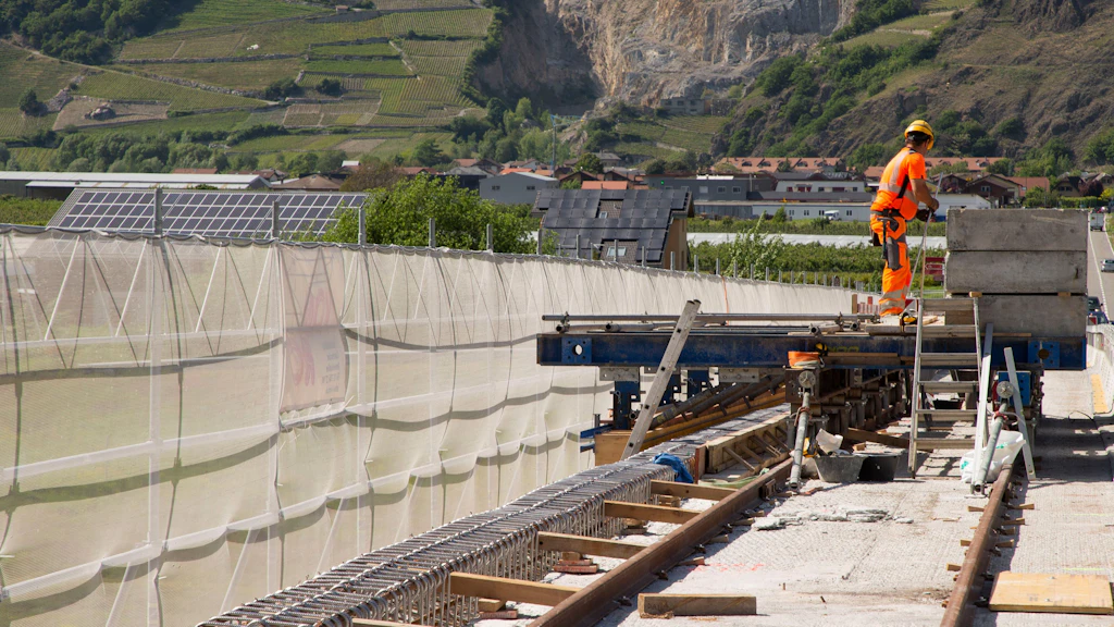 Ouvrier en tenue de sécurité orange debout sur un pont d'une route avec des filets de protection et un paysage de collines en arrière-plan