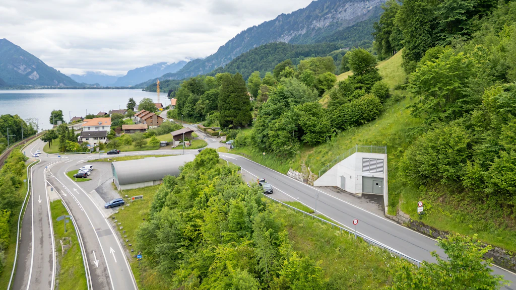 Strasse mit Tunnelportal am Hang neben einem See und bewaldeten Bergen im Hintergrund.