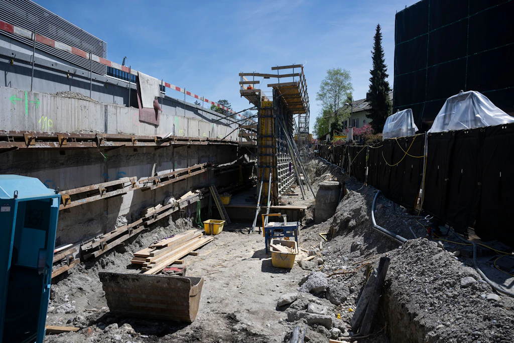 Baustelle mit einer tiefen Baugrube, die von Betonwänden und Holzverschalungen eingefasst ist. Im Hintergrund steht ein gelbes Gerüst mit einer Treppe, das von Bauarbeitern genutzt werden kann. Links sind Bauholz, Werkzeuge und ein blauer Container zu sehen. Rechts begrenzt ein schwarzer Bauzaun das Gelände, dahinter stehen Bäume und Gebäude. Die Szene zeigt zahlreiche Baumaterialien und Kabel auf dem Boden.