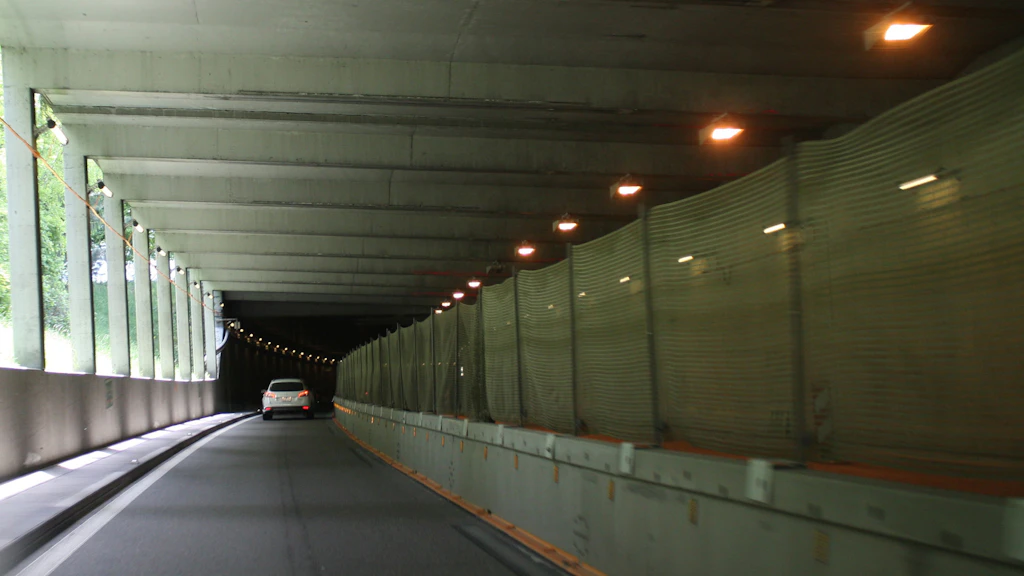 Beleuchteter Strassentunnel mit einer Fahrzeugspur und einem Auto auf der linken Fahrspur. Die rechte Spur ist abgetrennt mit rechteckigen Metallen mit einem Zaun obendrauf.