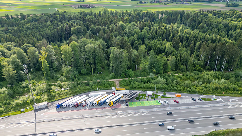 Luftaufnahme eines Rastplatzes mit parkenden Lastwagen neben einer Autobahn und einem dichten Wald im Hintergrund