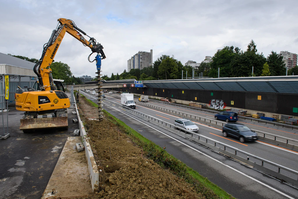 Eine gelbe Baumaschine mit einem Bohraufsatz steht auf einer Baustelle erhöht neben der Autobahn. Die Maschine bohrt Löcher in den Boden entlang einer Betonabsperrung. Auf der Strasse fahren mehrere Fahrzeuge. Im Hintergrund sind ein langgestrecktes Gebäude mit dunkler Fassade, hohe Wohnhäuser und zahlreiche Bäume zu sehen. Der Himmel ist bewölkt.