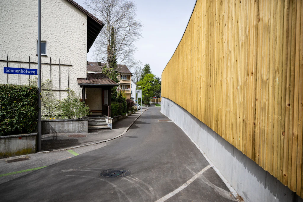 Eine hohe, durchgehende Lärmschutzwand aus senkrechten Holzbrettern verläuft entlang einer asphaltierten Strasse. Links stehen Wohnhäuser mit hellen Fassaden und dunklen Dächern. Ein blaues Strassenschild mit weißer Schrift trägt die Aufschrift "Sonnenhofweg". Im Hintergrund sind weitere Häuser und Bäume zu sehen. Der Himmel ist bedeckt.