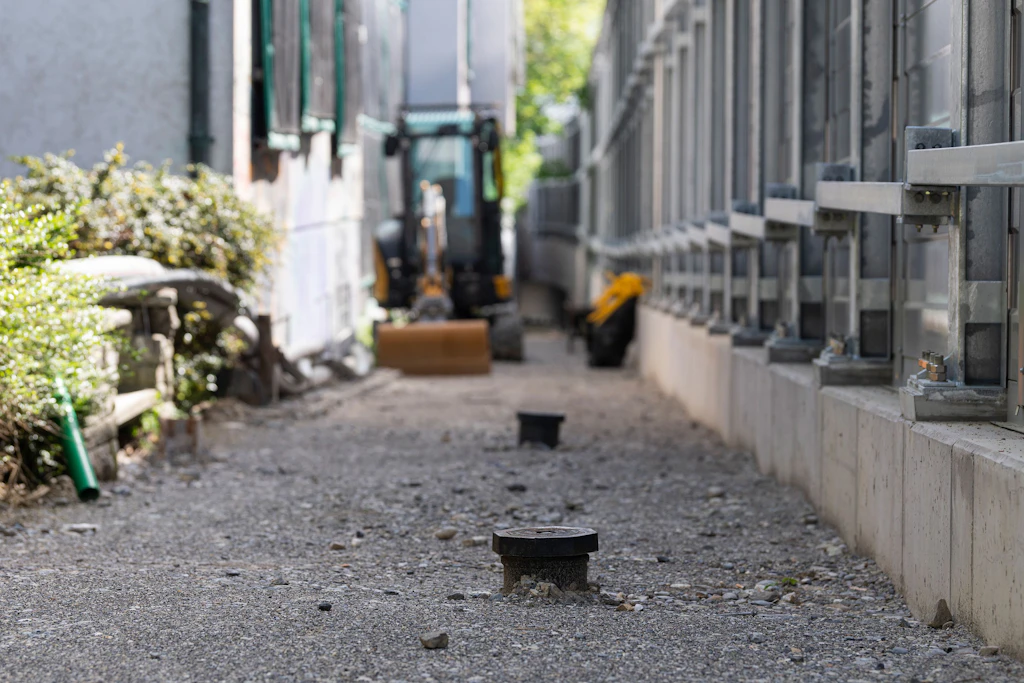 Im Vordergrund verläuft ein unbefestigter Weg mit zwei runden Abdeckungen, die aus dem Boden ragen. Rechts ist die Rückseite einer Lärmschutzwand aus Metall zu sehen, die auf einer Betonmauer steht. Links ist eine Gebäudefassade mit Fenstern zu sehen, die von schwarzen, lärmdämmenden Matten zugedeckt sind. Im Hintergrund steht ein kleiner Bagger mit Schaufel auf dem Weg. Die Szene ist von Tageslicht erhellt.