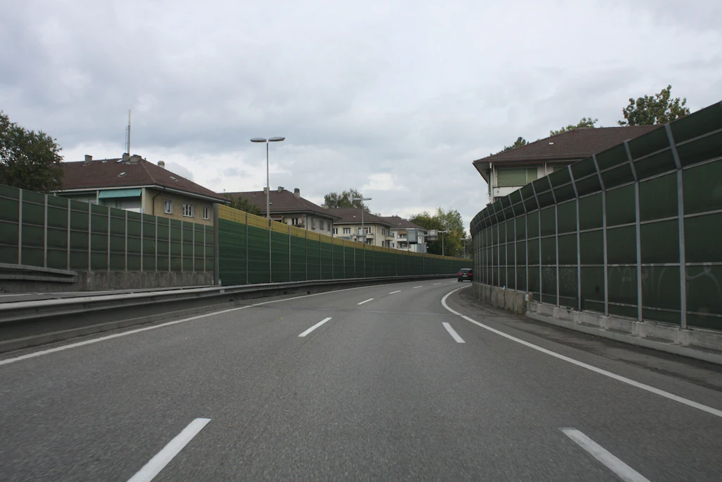 Eine Autobahn mit mehreren Fahrspuren verläuft zwischen hohen, grünen Lärmschutzwänden. Die Wände bestehen aus Metallrahmen mit Gitterstruktur und sind beidseitig entlang der Fahrbahn aufgestellt. Im Hintergrund stehen Wohnhäuser mit dunklen Dächern und hellen Fassaden. Der Himmel ist bewölkt, die Beleuchtung wirkt gleichmäßig und diffus. Ein Strassenlaternenmast steht am linken Rand der Fahrbahn.