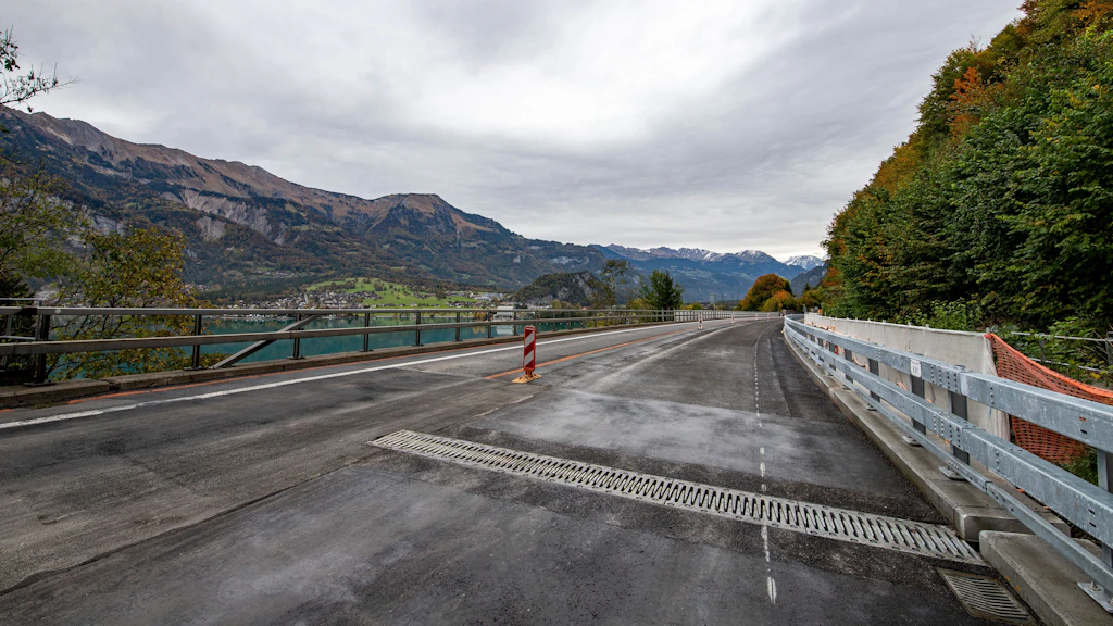 Leere asphaltierte Strasse mit Leitplanke und Baustellenabsperrung vor Berglandschaft und See