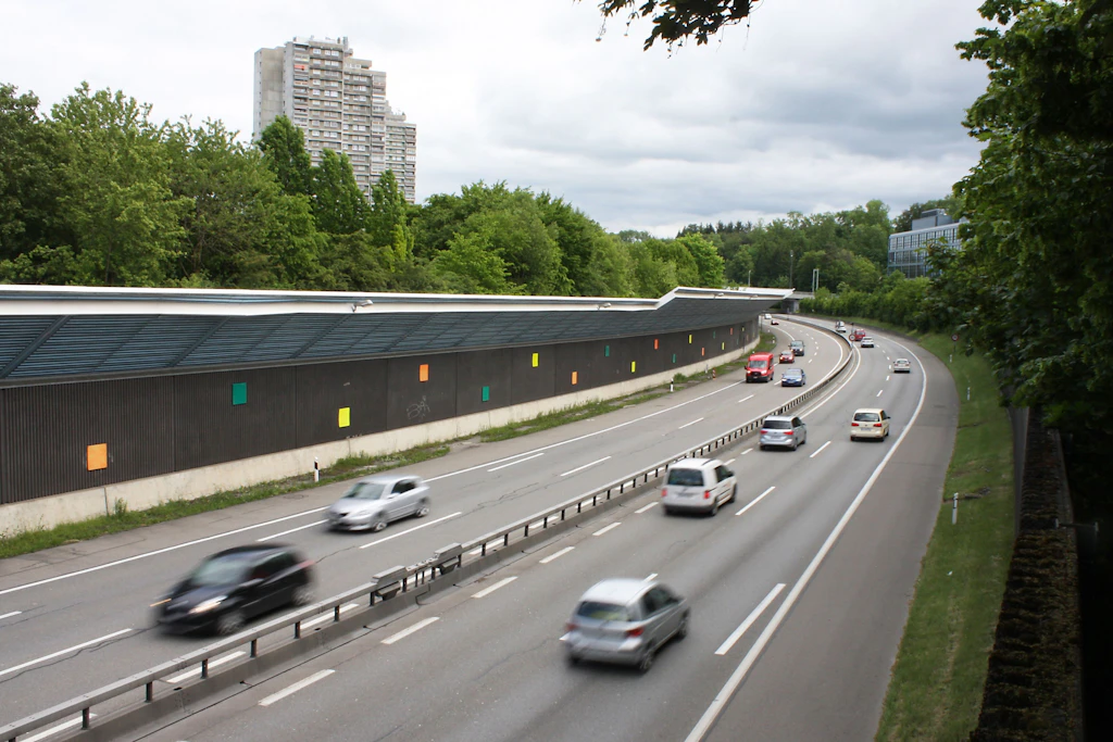 Eine Autobahn verläuft durch eine grüne Umgebung. Auf der linken Seite steht eine lange Lärmschutzwand mit quadratischen, farbigen Elementen. Im Hintergrund sind hohe Wohngebäude und dichter Baumbestand zu sehen. Mehrere Fahrzeuge bewegen sich in beide Richtungen. Der Himmel ist bewölkt.