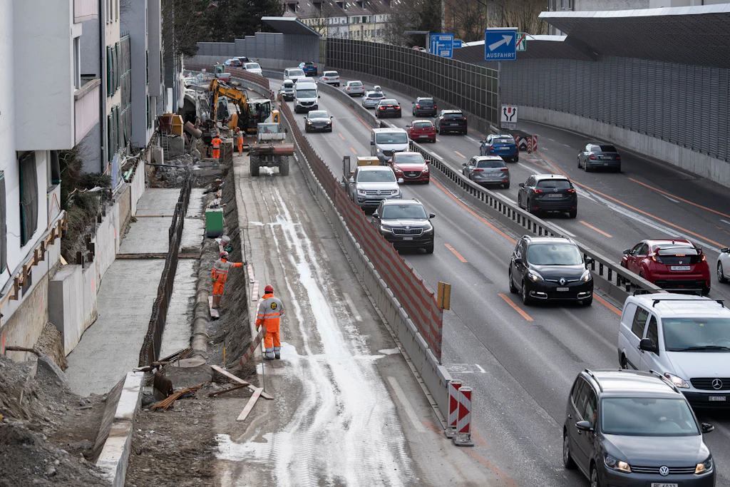 Mehrspurige Autobahn mit dichtem Verkehr. Auf der linken Seite arbeiten mehrere Personen in orangefarbener Schutzkleidung an einer Baustelle, die durch Absperrungen vom Strassenbereich getrennt ist. Ein Bagger steht im Baustellenbereich. Die Fahrbahn ist durch eine Betonbarriere und rot-weiße Markierungen von der Baustelle abgegrenzt. Im Hintergrund sind Gebäude und eine Lärmschutzwand sichtbar. Über der Strasse befinden sich blaue Wegweiser mit weißer Schrift.