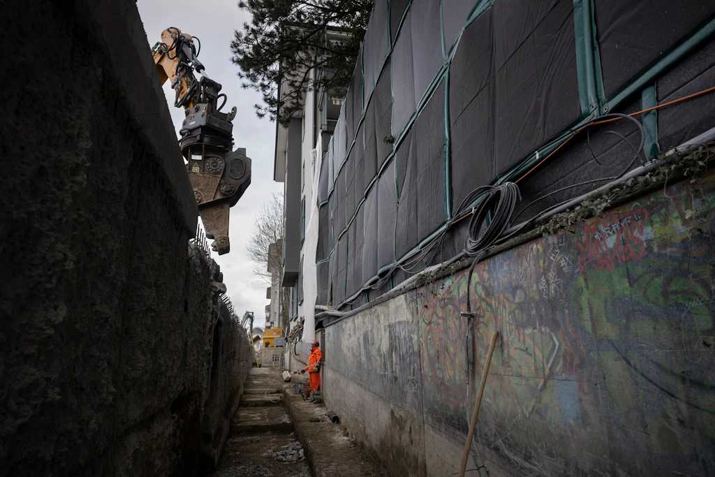 Eine Baustelle mit einer tiefen, schmalen Grube, die sich entlang einer Wand erstreckt. Links ist ein großer Baggerarm mit Greifvorrichtung zu sehen, der über die Grube ragt. Rechts, nur etwa zwei Meter entfernt, verläuft die Fassade von Wohnhäusern. Die Fassade ist mit schwarzen, lärmdämmenden Matten abgedeckt. Im Hintergrund steht eine Person in orangefarbener Arbeitskleidung am Rand der Grube. Oben wachsen Äste eines Baumes über die Baustelle.