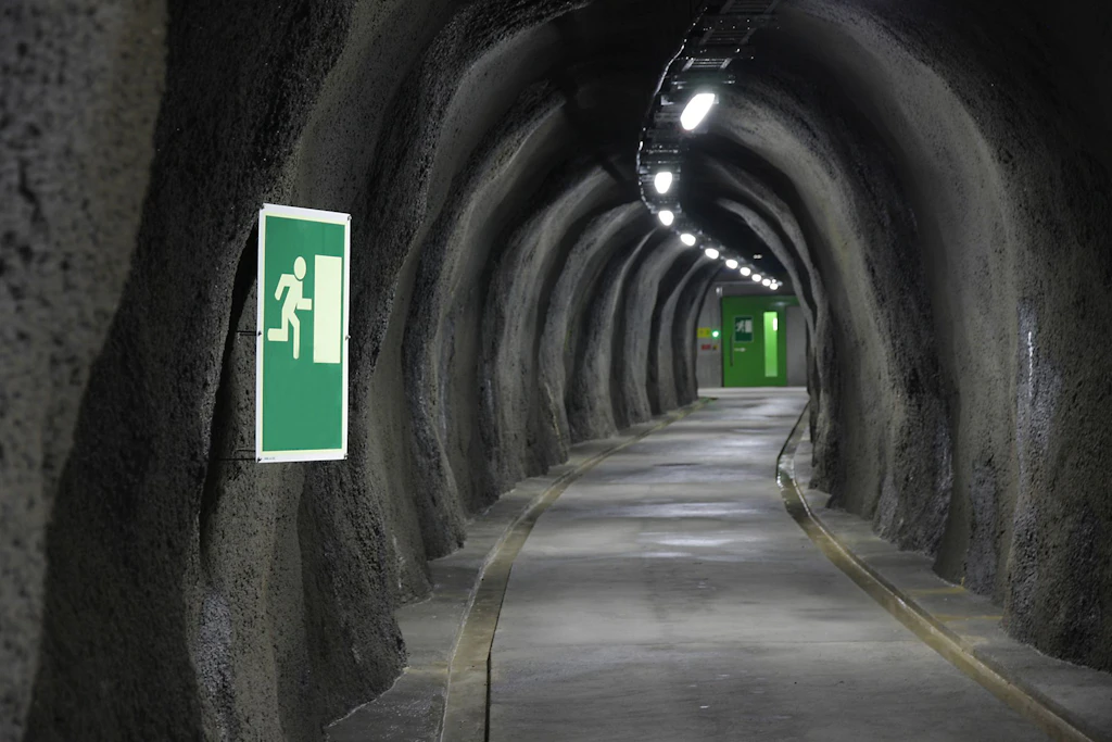 Tunnel für Fussgänger mit unregelmäßigen Felswänden, beleuchtetem Fluchtwegschild und grüner Ausgangstür am Ende.