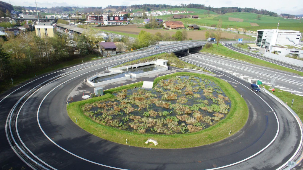Luftaufnahme einer Kurve einer Autobahnausfahrt mit einem runden Wasserbecken, das mit Schilf bewachsen ist.