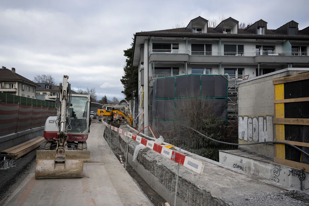 Eine Baustelle mit mehreren Baumaschinen und Absperrungen entlang der Autobahn beim Ostring. Links steht ein Bagger mit breiter Schaufel, dahinter sind weitere Baumaschinen und Baumaterialien zu sehen. Rechts befindet sich ein mehrstöckiges Wohngebäude mit grauer Fassade, davor ein rundes Baugerüst schwarzen, lärmdämmenden Matten. Ein rechteckiger Betonbau mit Graffiti und Holzverkleidung grenzt an die Strasse. Der Himmel ist bewölkt.