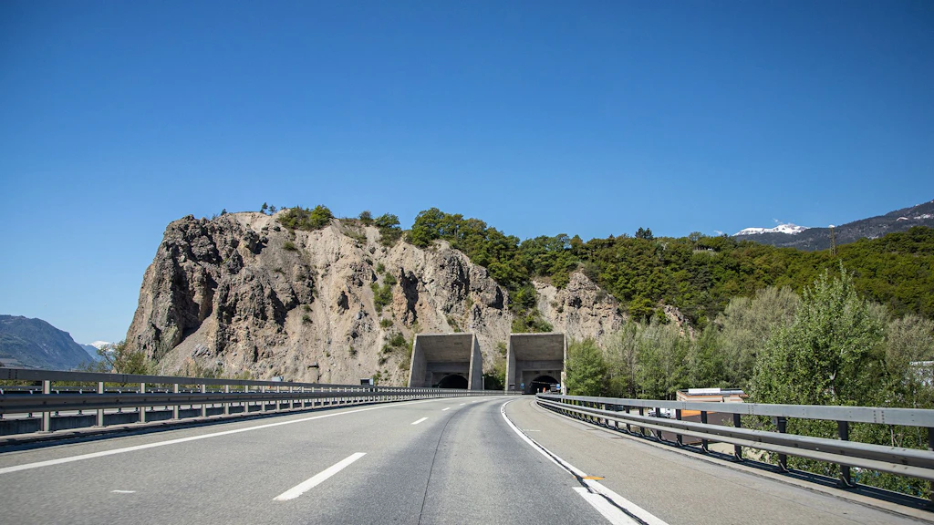Autobahn mit zwei Tunnelportalen des Tunnel Sierre, die in einen bewaldeten Felsen führen, unter blauem Himmel.
