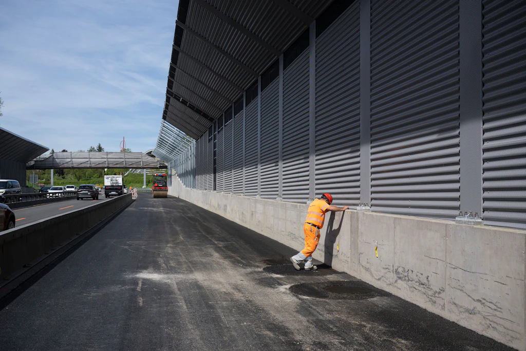 Eine Person in orangefarbener Arbeitskleidung und rotem Helm steht auf frisch asphaltiertem Strassenbelag und lehnt sich mit beiden Händen gegen eine hohe, graue Lärmschutzwand. Die Wand besteht aus horizontalen Lamellen und ist auf einem Betonfundament montiert. Links im Bild verläuft die mehrspurige Autobahn mit mehreren Fahrzeugen. Sie ist mit Betonelementen von der Baustelle abgetrennt. Der Himmel ist klar und blau.