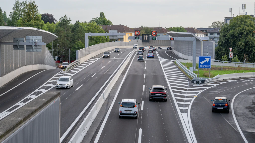 Mehrspurige Autobahn beim Anschluss Ostring, auf dern verschiedene Autos fahren. Links und rechts sind hohe Lärmschutzwände zu sehen. Im Hintergrund stehen Wohnhäuser und Bäume. Über der Fahrbahn befinden sich elektronische Verkehrszeichen und eine grosse Brücke. Rechts ist ein blaues Schild mit dem Wort "Ausfahrt" deutlich lesbar.