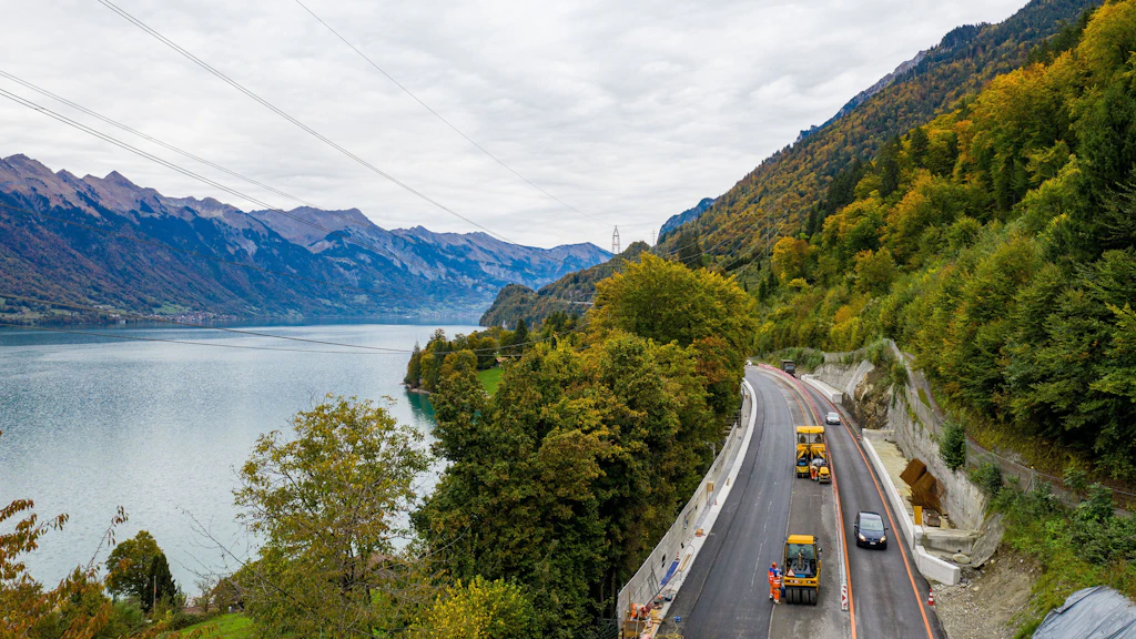 Blick auf einen See auf der linken Seite mit Bergen im Hintergrund und eine Strasse auf der rechten Seite mit mehreren Baumaschinen.