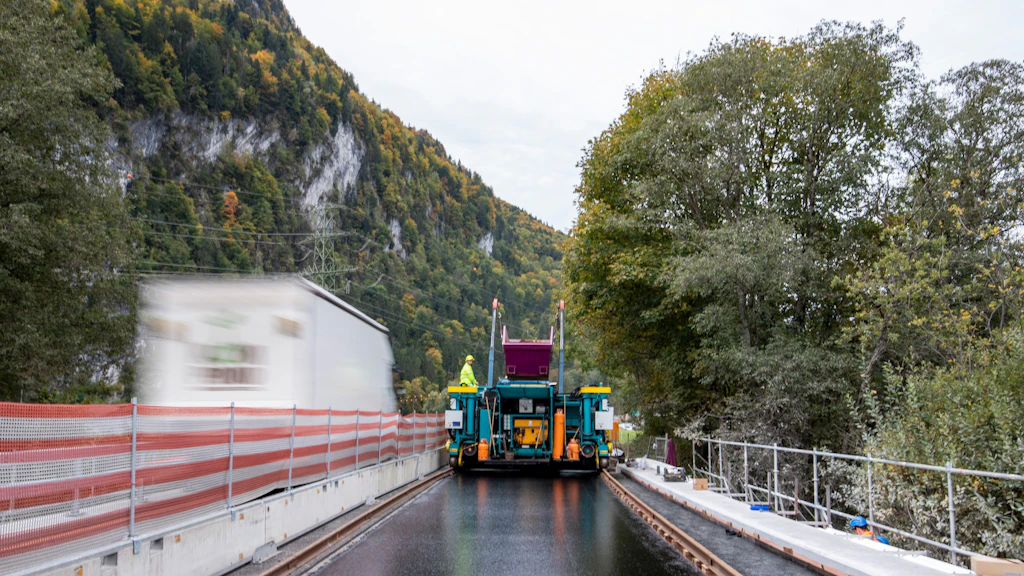 Grosses Gerät zum Belagseinbau auf einer Fahrspur auf zwei Schienen. Linke Spur mit Betonelementen und Sichtschutzzaun vom Verkehr abgetrennt.