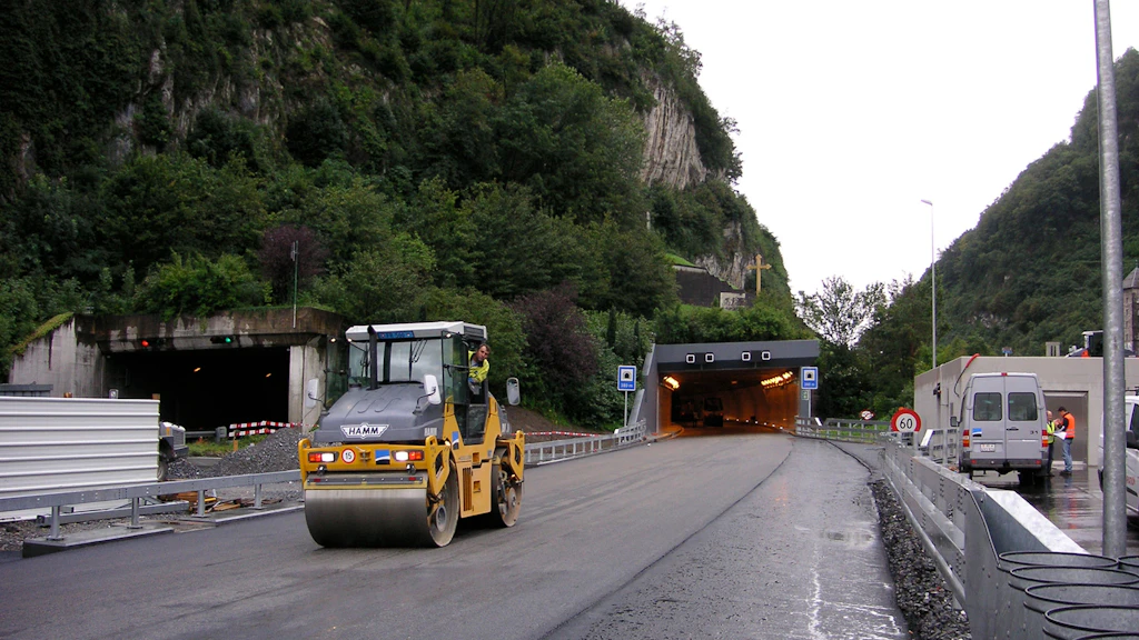 Strassenwalze auf nasser Strasse vor zwei Tunnelportalen in bergiger Landschaft mit Baustellenpersonal