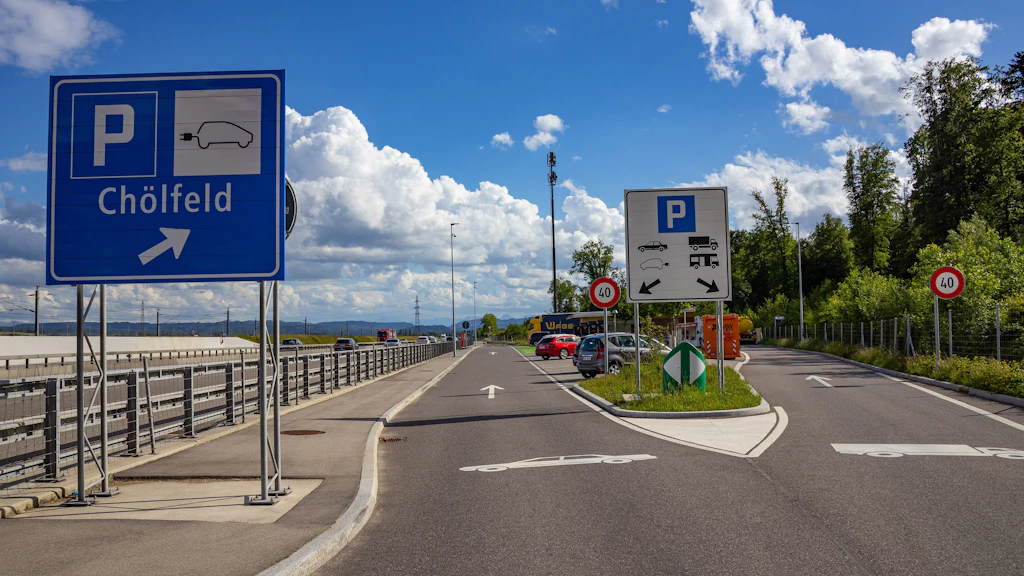 Strasse mit Verkehrsschildern für Parkplatz Chölfeld und Rastplatz mit Lkw- und Pkw-Symbolen unter blauem Himmel mit Wolken