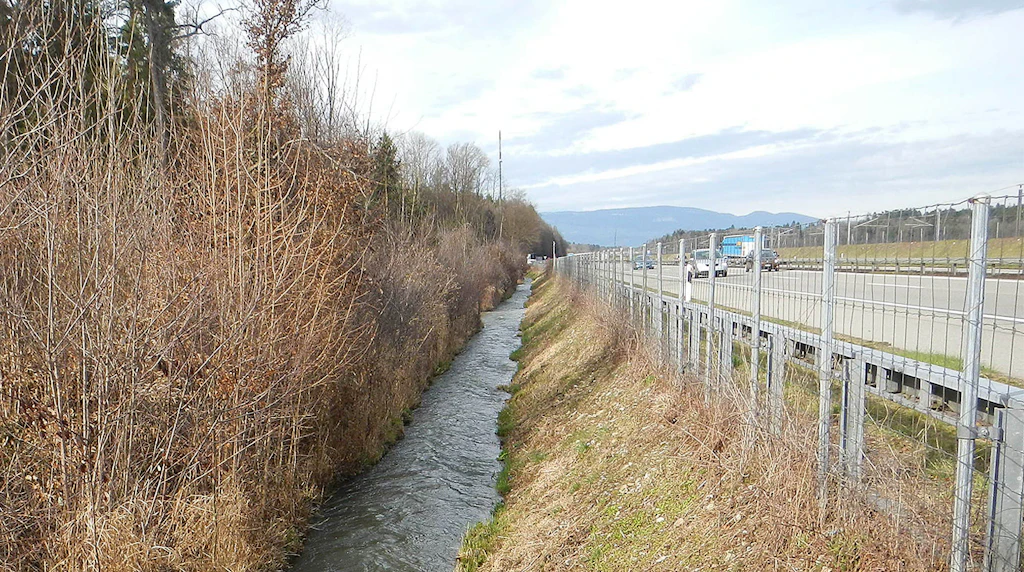 Kanalisierter Bach mit kahlen Bäumen links und Autobahn mit Leitplanke rechts unter bewölktem Himmel