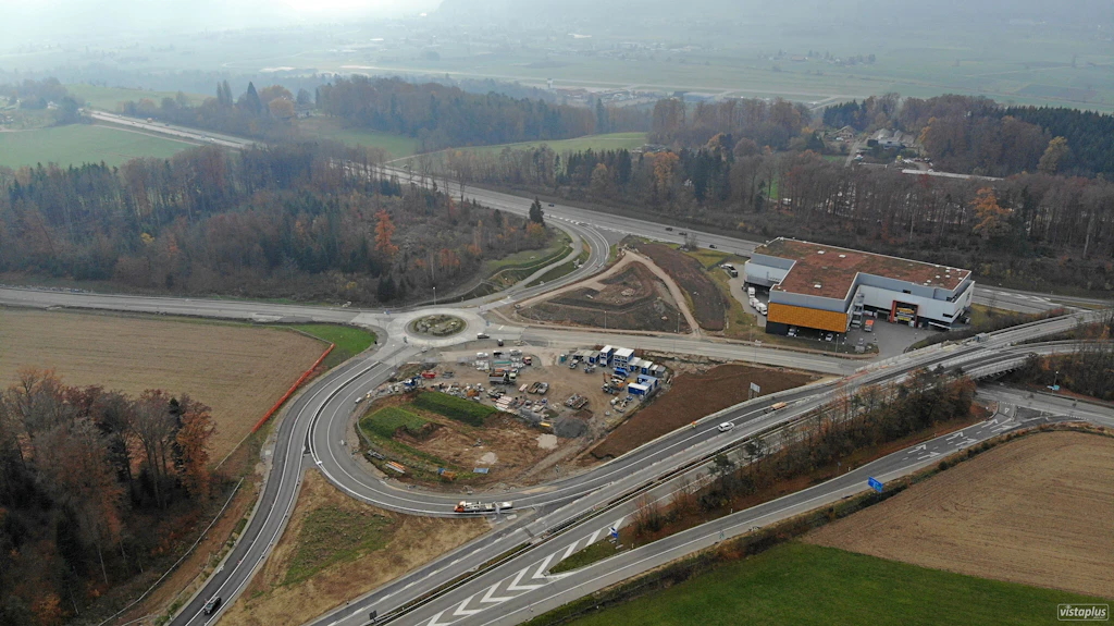Luftaufnahme einer Autobahnanschlussstelle mit Kreisverkehr im Bau, angrenzenden Feldern und einem Gebäude mit rotem Dach