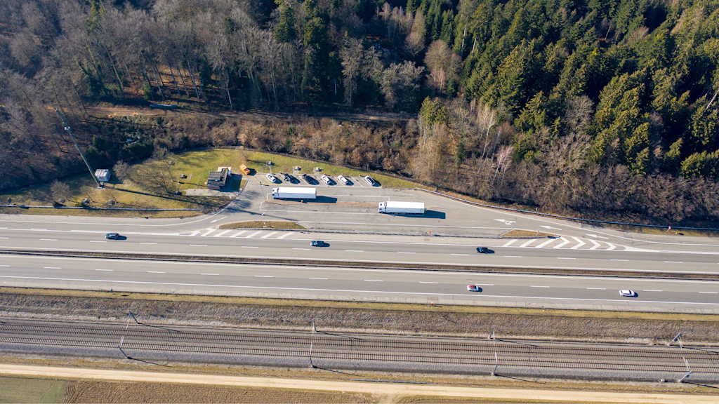 Luftaufnahme eines Rastplatzes neben einer Autobahn mit parkenden Fahrzeugen und einem angrenzenden Waldgebiet