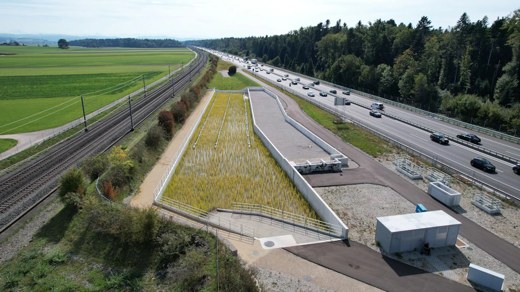 Luftaufnahme einer Autobahn mit mehreren Fahrspuren neben einer Bahnstrecke Dazwischen ein grosses, rechteckiges Wasserbecken mit Schilf und einem zweiten, leeren Becken daneben.