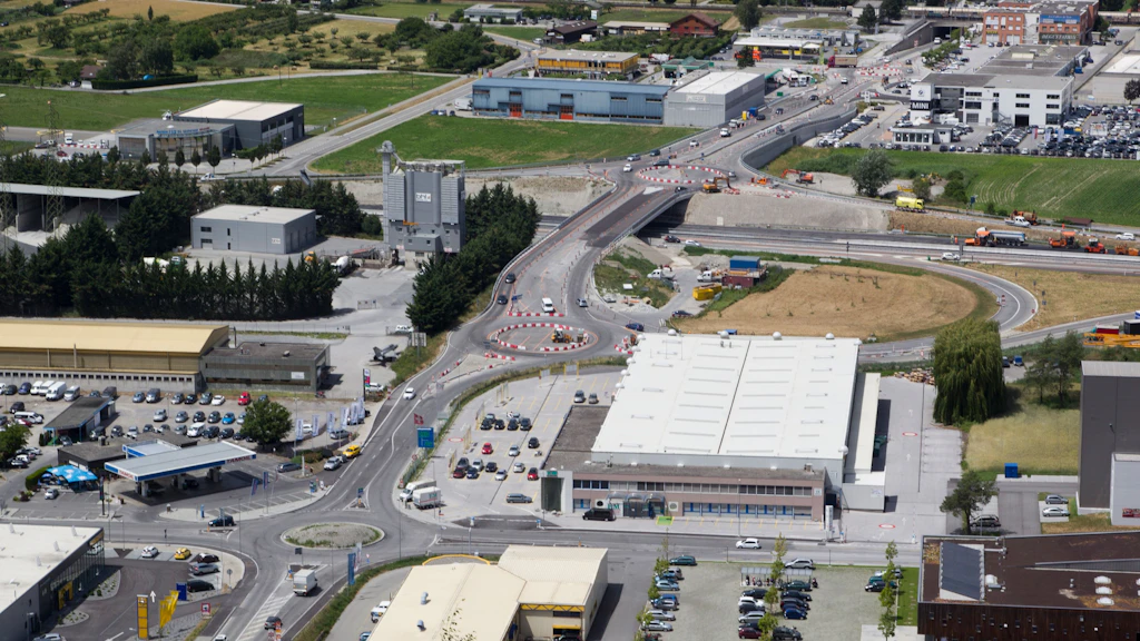 Luftaufnahme eines Industriegebiets mit mehreren Gebäuden und Fahrzeugen. In der Bildmitte eine Strasse über die Autobahn mit Kreisel im Bau auf beiden Seiten der Autobahn