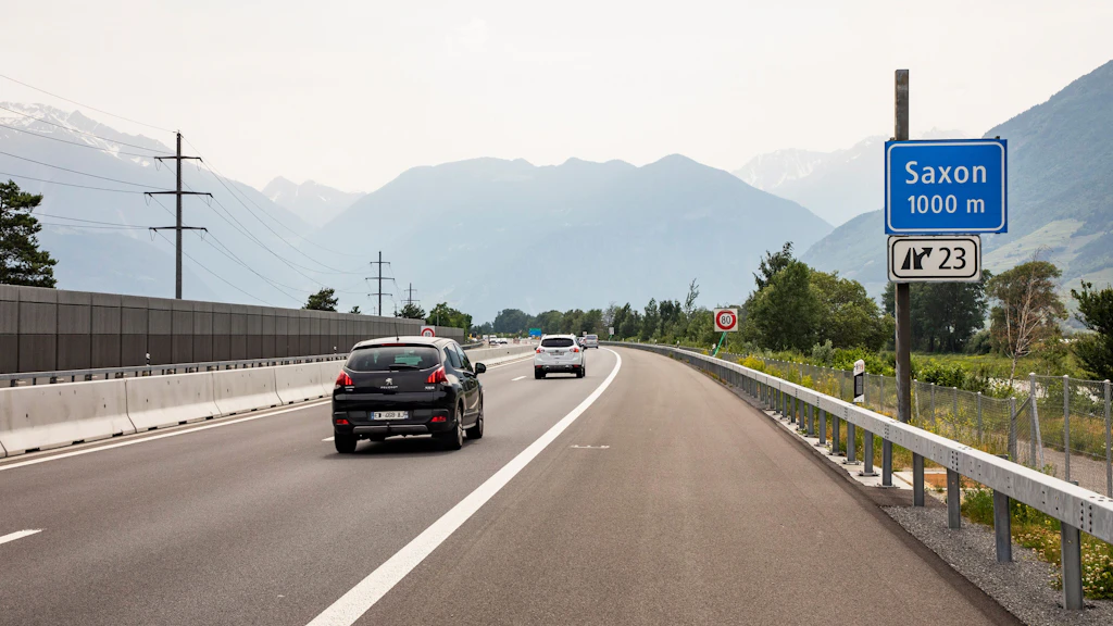 Autoroute avec plusieurs voitures, panneau indiquant Saxon à 1000 mètres, paroi antibruit à gauche et montagnes en arrière-plan