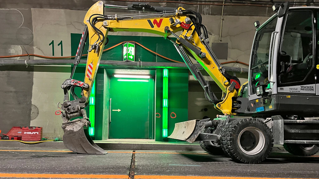 Une pelleteuse à flèche jaune se trouve dans un tunnel éclairé, devant une porte de secours verte