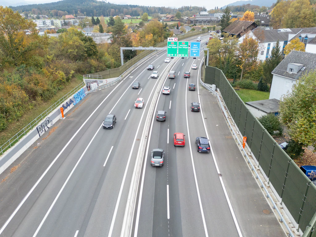 Eine Autobahn mit mehreren Fahrspuren verläuft durch eine grüne Landschaft. Auf beiden Seiten der Fahrbahn stehen hohe Lärmschutzwände aus Metall oder Kunststoff mit schallabsorbierenden Elementen. Mehrere Fahrzeuge fahren in beide Richtungen. Im Hintergrund befinden sich Wohnhäuser, Bäume mit herbstlicher Färbung und ein bewaldeter Hügel. Über der Fahrbahn hängen grüne Hinweisschilder mit weisser Schrift.