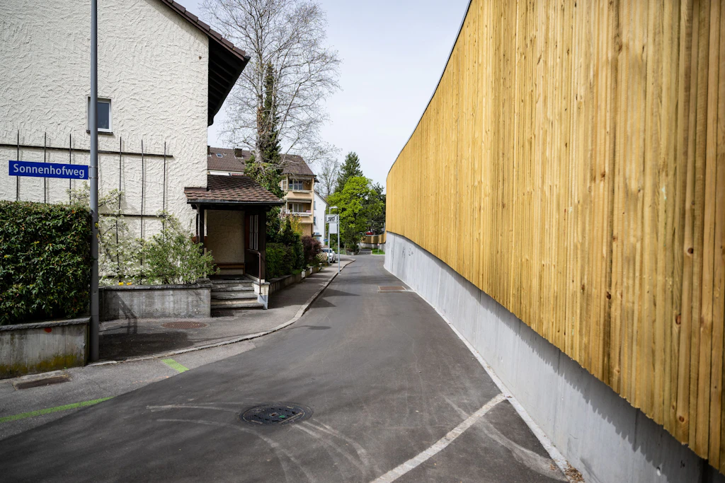 Eine hohe, durchgehende Lärmschutzwand aus senkrechten Holzbrettern verläuft entlang einer asphaltierten Strasse. Links stehen Wohnhäuser mit hellen Fassaden und dunklen Dächern. Ein blaues Strassenschild mit weißer Schrift trägt die Aufschrift "Sonnenhofweg". Im Hintergrund sind weitere Häuser und Bäume zu sehen. Der Himmel ist bedeckt.
