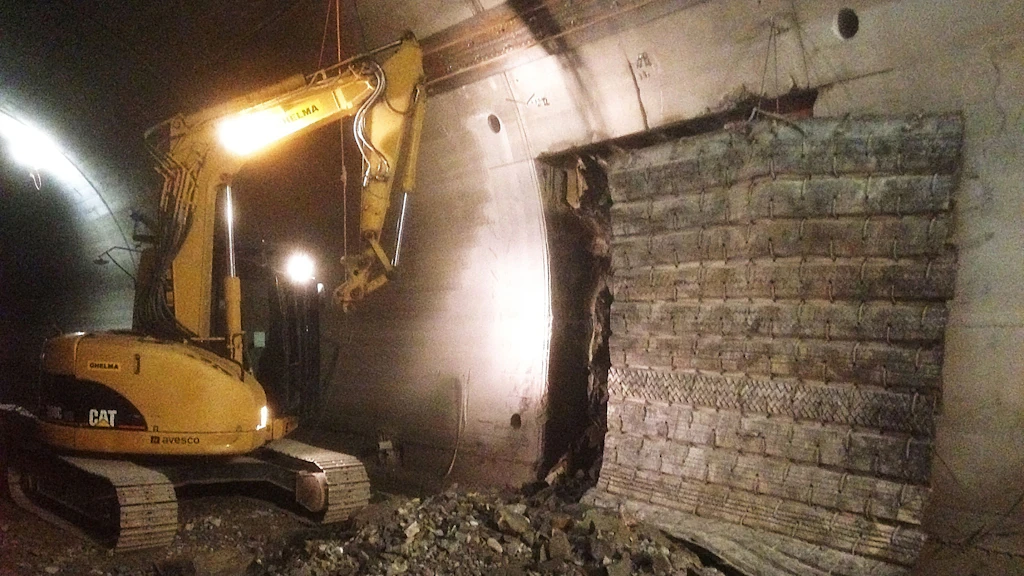 Une pelleteuse équipée d'un système d'éclairage travaille dans un tunnel sur une ouverture pratiquée dans la paroi, à côté d'une paroi de tunnel revêtue de pierres