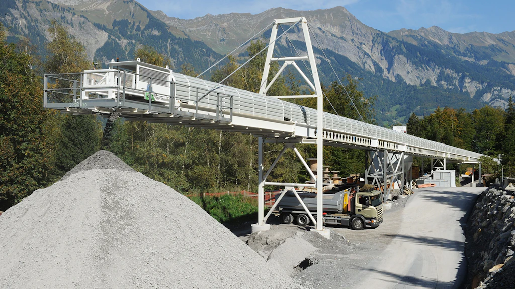 Longue installation de convoyeurs au-dessus d'un chantier avec des tas de gravier, sur fond de montagnes