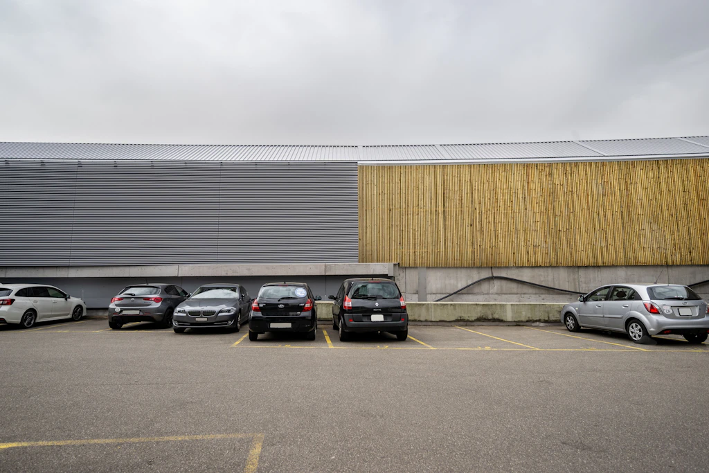Mehrere Autos stehen nebeneinander auf einem asphaltierten Parkplatz. Im Hintergrund verläuft eine hohe Lärmschutzwand, die aus zwei unterschiedlichen Abschnitten besteht: Links sind horizontale Metalllamellen zu sehen, rechts vertikale Holzbalken. Der Himmel ist bewölkt.