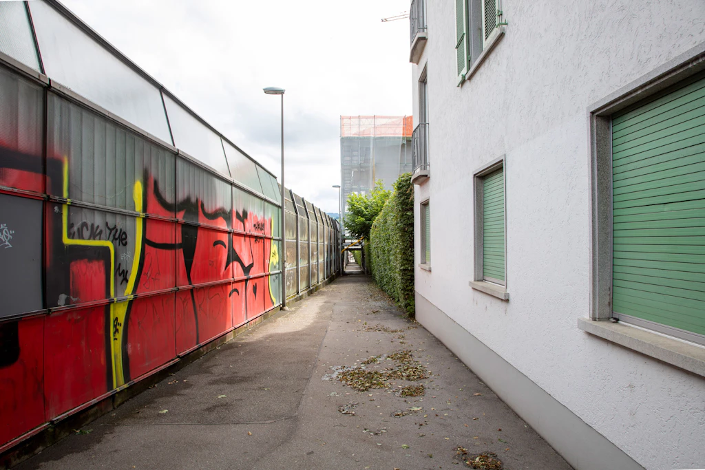 Ein schmaler Durchgang verläuft zwischen einer grauen Metallwand mit grossflächigen roten und gelben Graffiti und einer weißen Hausfassade mit grünen Fensterläden. Der Boden ist asphaltiert und teilweise mit Laub bedeckt. Am Ende des Durchgangs sind ein weiteres Gebäude und eine Straßenlaterne sichtbar. Die Szene ist von Tageslicht erhellt.