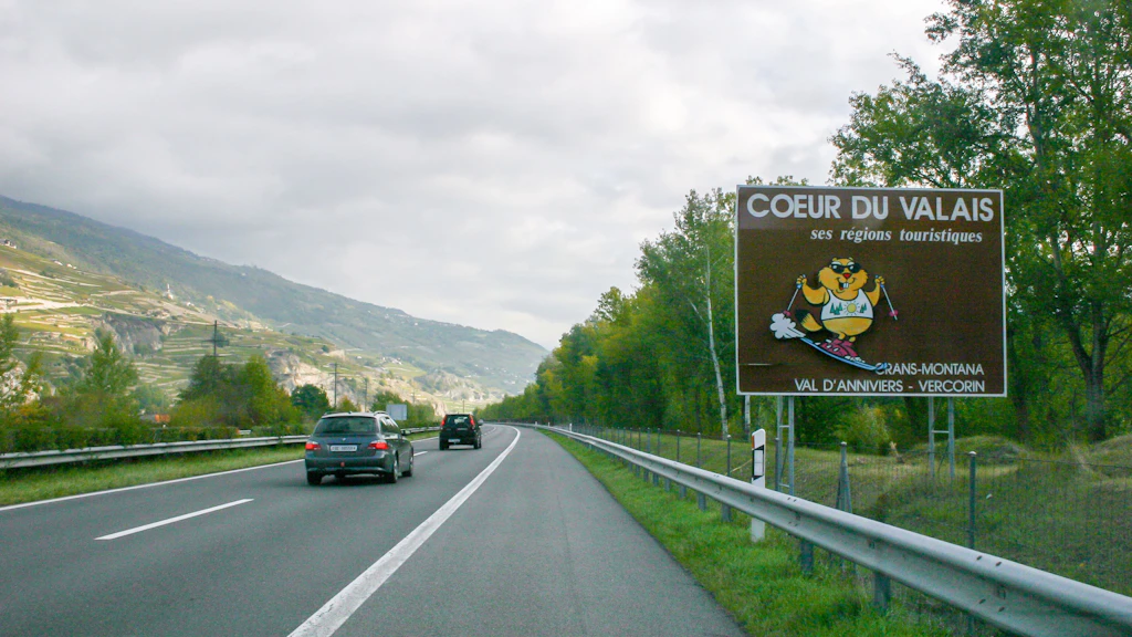 Une autoroute à plusieurs voies traverse un paysage verdoyant et légèrement vallonné, parsemé d'arbres et de vignobles. À gauche, plusieurs voitures roulent en direction d'une vallée entourée de montagnes. À droite se trouve un grand panneau indicateur marron avec une inscription blanche et une illustration colorée. Le panneau montre un personnage jaune coiffé d'un chapeau et tenant des raisins, et répertorie les régions touristiques. Le ciel est nuageux.
