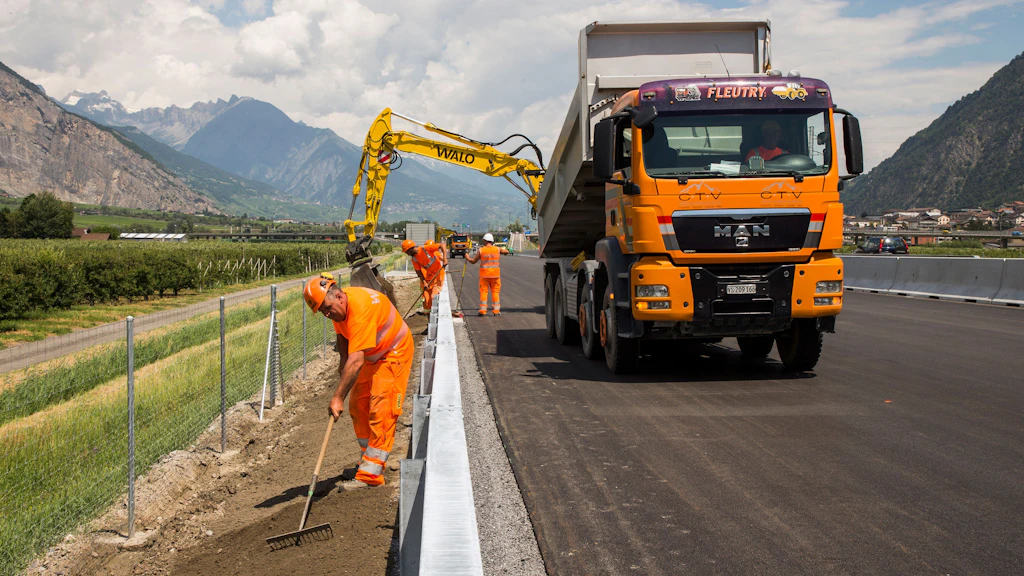 Travailleurs en tenue orange posant une clôture le long d'une route asphaltée avec un camion orange à l'arrêt et des montagnes en arrière-plan