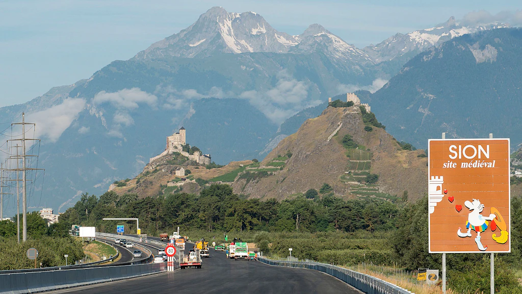 Une autoroute avec des véhicules devant des collines boisées et les ruines d'un château, avec de hautes montagnes en arrière-plan ; à droite, un panneau marron portant l'inscription « SION site médiéval » et le dessin d'un personnage avec un cœur.