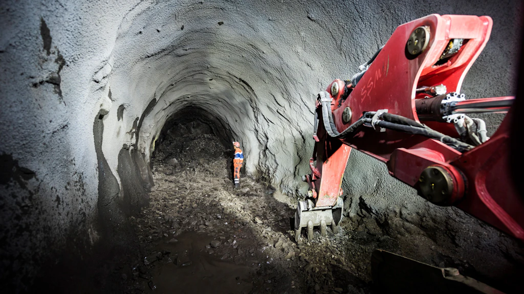 Tunnel voûté en cours de construction, avec un bras de pelleteuse rouge au premier plan et un ouvrier du bâtiment à l'arrière-plan.