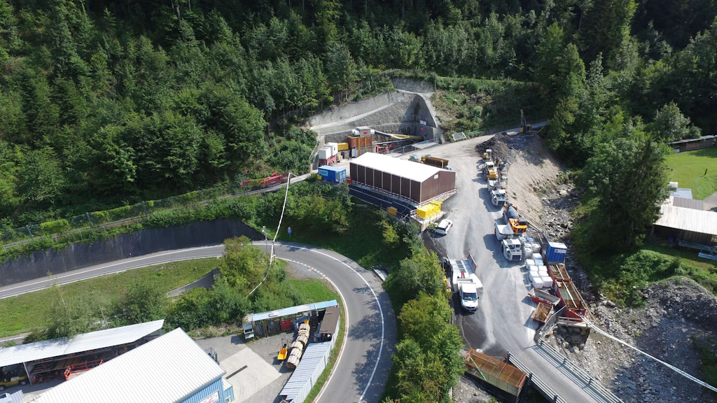 Vue aérienne d'un chantier avec plusieurs véhicules et conteneurs à l'entrée est d'un tunnel, dans un paysage montagneux boisé.