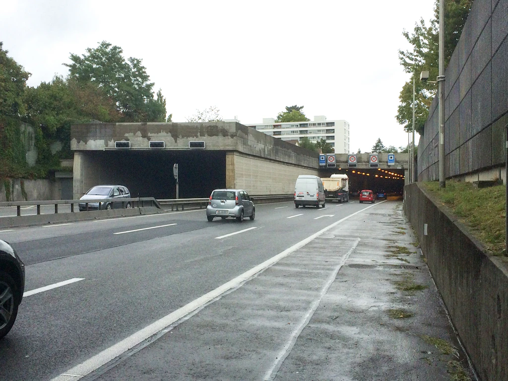 Zwei Tunnelportale mit rechteckigen Öffnungen liegen nebeneinander an der Autobahn. Mehrere Fahrzeuge sind auf der Strasse. Über dem rechten Tunnel sind Verkehrszeichen und elektronische Anzeigen angebracht. Die Fahrbahn ist nass, der Himmel bedeckt. Links und rechts der Strasse stehen Betonwände, im Hintergrund sind Bäume und ein mehrstöckiges Wohngebäude sichtbar.
