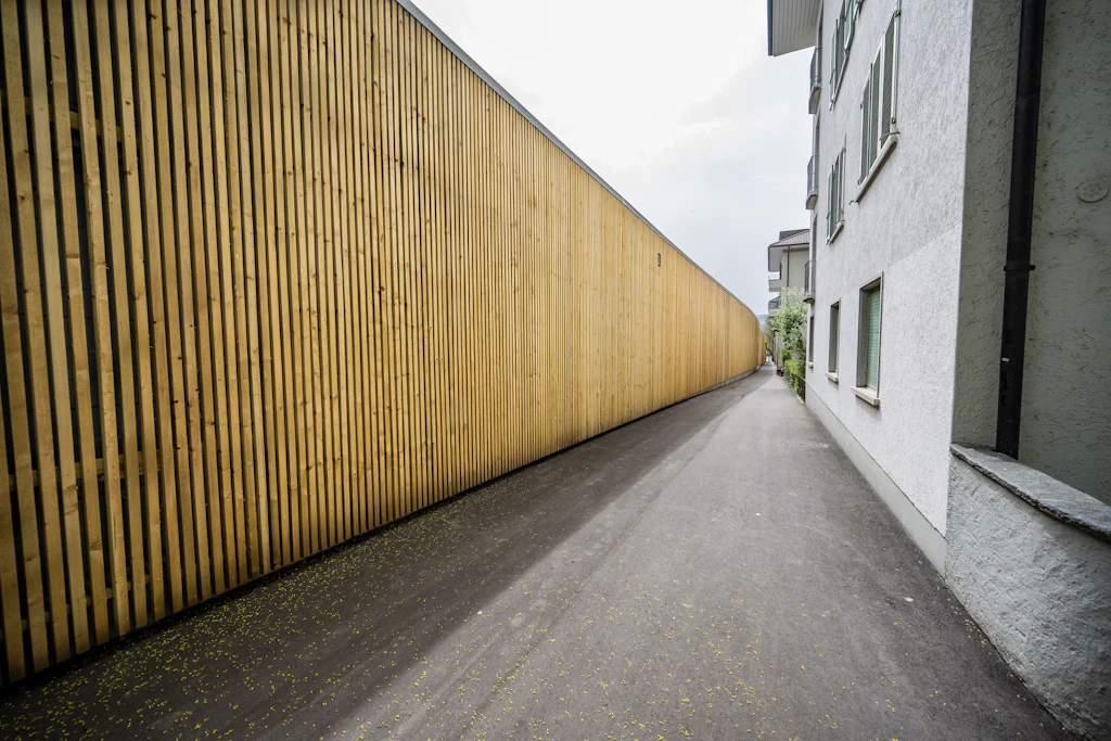 Eine hohe Lärmschutzwand aus senkrechten, hellen Holzbrettern verläuft entlang eines asphaltierten Gehweges. Rechts steht ein mehrstöckiges, helles Wohnhaus mit rauer Fassade und Fenstern. Die Wand und das Gebäude bilden einen schmalen, geradlinigen Durchgang, der in die Tiefe führt. Der Boden ist sauber, und es sind keine Menschen oder Fahrzeuge zu sehen.