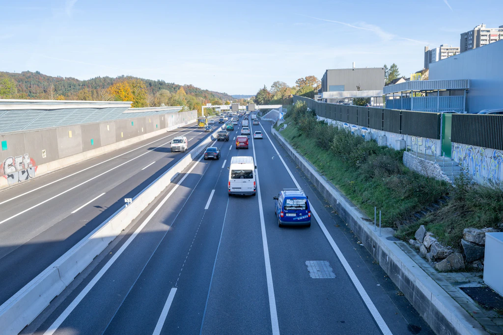 Mehrspurige Autobahn mit mittlerem Verkehrsaufkommen. Links und rechts der Fahrbahn verlaufen hohe Lärmschutzwände, rechts zusätzlich ein bepflanzter Hang und moderne Gebäude im Hintergrund. Die Szene zeigt herbstliche Bäume und einen klaren Himmel.