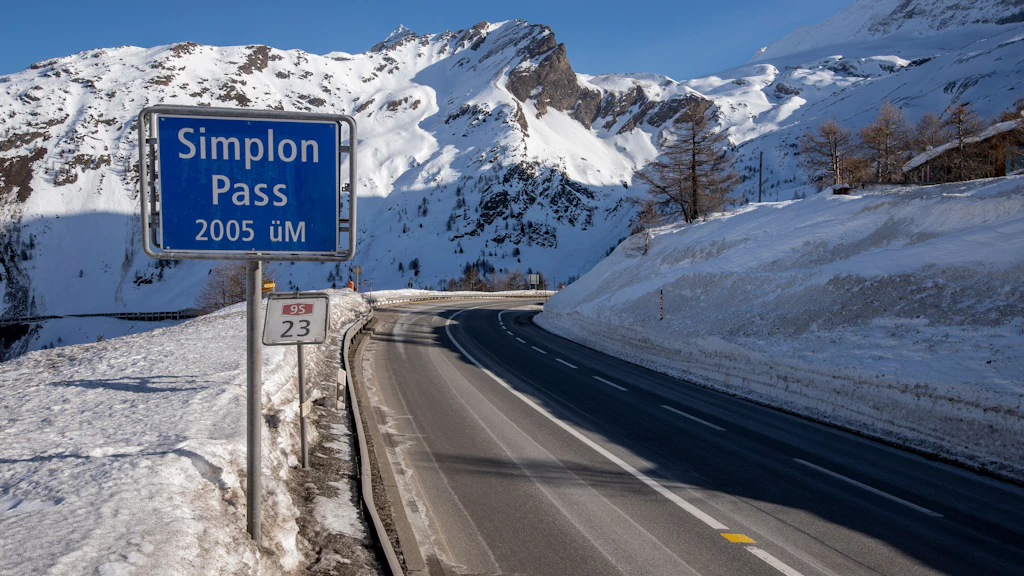 Route parsemée de restes de neige, avec un panneau de signalisation bleu indiquant « Col du Simplon 2005 m d'altitude » devant des montagnes enneigées