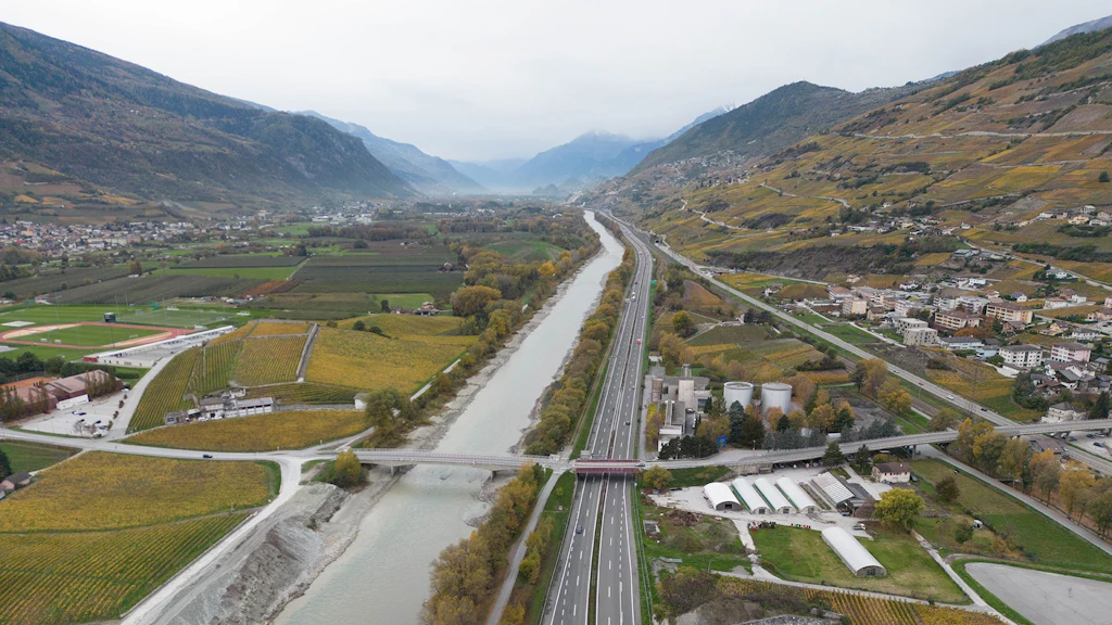 Vue aérienne de l'autoroute A9 entre Sion et Sierre. L'autoroute passe au centre de l'image, parallèlement au Rhône. À gauche et à droite, on aperçoit des champs et des habitations, avec une chaîne de montagnes en arrière-plan.